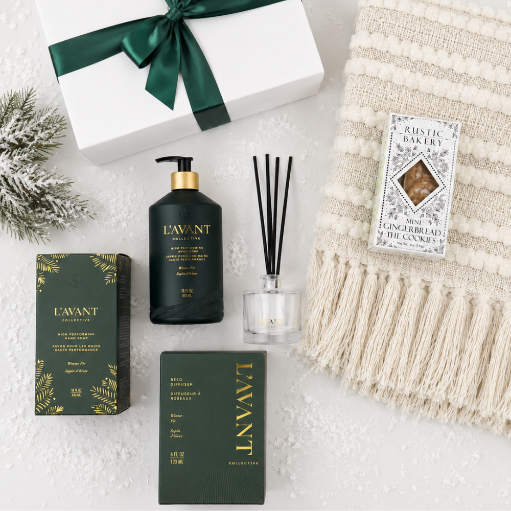 A curated gift set featuring L'AVANT products, including a dark green gift box with a green ribbon, a black body mist bottle with a gold cap, a reed diffuser with black reeds, and a dark green box, arranged on a white textured surface next to a cream knit blanket and a small evergreen sprig.