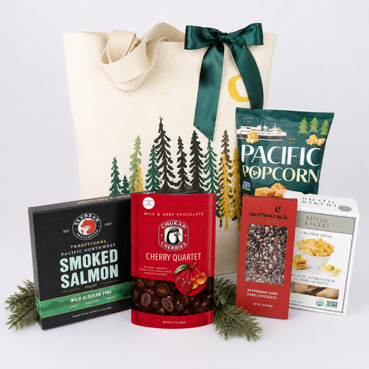 A gift basket set featuring a cream canvas tote bag with a green ribbon and pine tree print, surrounded by assorted snack boxes and bags including Trader Joe's Smoked Salmon, G. Maxwell's Cherry Quartet, and Pacific Popcorn.