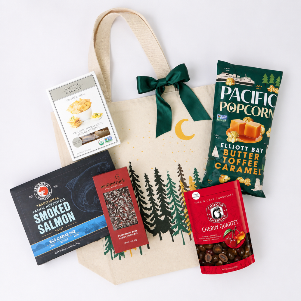 A cream canvas gift tote bag with a dark green satin bow and printed pine trees and moon, surrounded by gourmet snacks including Pacific Popcorn, Smoked Salmon, and Cherry Quartermelon on a white background.
